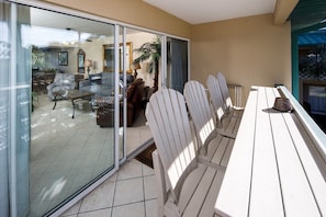 Um pátio iluminado pelo sol, com mesa e cadeiras de madeira, porta de vidro deslizante que dá para uma sala interna e vista para uma palmeira.