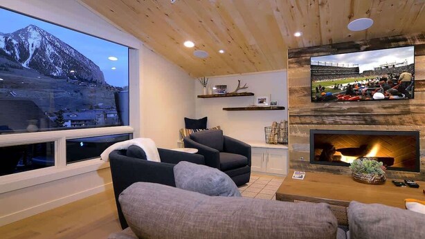 Ein Wohnzimmer mit Kamin, einem an der Wand befestigten Fernseher und einem großen Fenster mit Blick auf einen Berg.