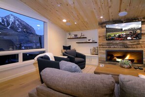 A living room with a fireplace, a television mounted on the wall, and a large window overlooking a mountain. - The Point (Mt. Crested Butte)