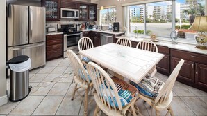 A modern kitchen with stainless steel appliances, a dining table with chairs, and large windows offering a view of the city. - Seaspray Condos - 101B (Fort Walton Beach)