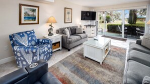 A living room with a sofa, armchair, coffee table, and a TV. There are framed pictures on the wall and a window with a view of the outdoors. - Seaspray Condos - 101B (Fort Walton Beach)