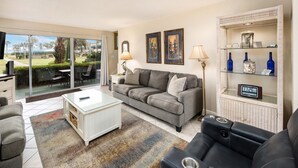 A living room with a grey sofa, a white coffee table, a black leather chair, and a glass-top table. There are two wall-mounted lamps, a clock, and a shelf with bottles and a decorative item. - Seaspray Condos - 101B (Fort Walton Beach)