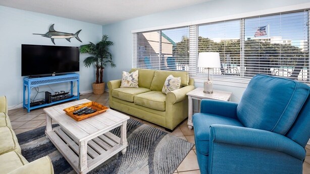 A living room with a television, sofa, armchair, coffee table, and a large window with blinds. - Seaspray Condos - 102C (Fort Walton Beach)