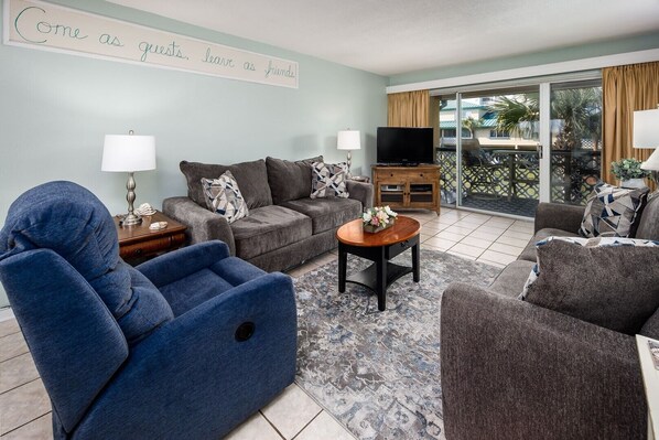 Un salon avec un canapé, un fauteuil et un téléviseur. Il y a un panneau sur le mur et on peut voir le balcon extérieur.