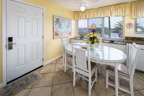 A dining area with a round glass table, white wicker chairs, a vase with yellow flowers, and a painting of a beach scene.