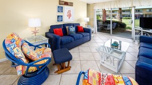 Una sala de estar con un sofá azul, sillones coloridos y una mesa de centro de vidrio. Hay cuadros enmarcados en la pared y un letrero que dice "CRAB SHACK".