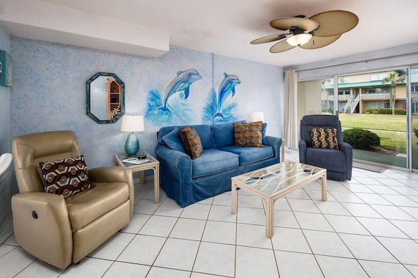 A living room with a blue sofa, a beige armchair, and a glass coffee table. There is a ceiling fan, a wall-mounted mirror, and a large window with a view of a building.