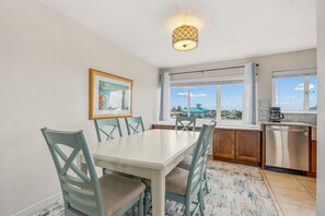 A dining area with a white table and chairs, a kitchen with a dishwasher, and a window with a view of the outside.