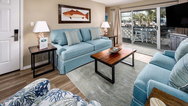 A living room with a sofa, a coffee table, and a television. There is a door and a window with a view of the outside.