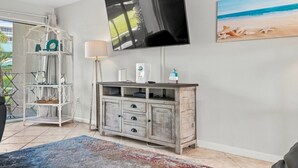 A living room with a flat-screen TV mounted on the wall, a wooden cabinet with drawers, a glass shelf unit, and a beach-themed painting. - Seaspray Condos - 109A (Fort Walton Beach)