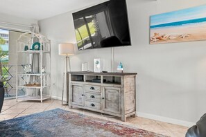 A living room with a flat-screen TV mounted on the wall, a wooden cabinet with drawers, a glass shelf unit, and a beach-themed painting. - Seaspray Condos - 109A (Fort Walton Beach)