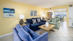 A living room with a blue sofa, a green armchair, and a wooden coffee table. There are two lamps, a painting of a beach scene, and a door with a sign.