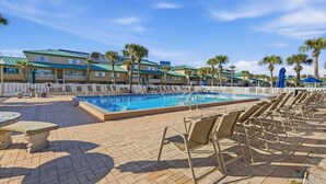 A pool area with lounge chairs, a pool, and a building with a green roof. - Seaspray Condos - 112B (Fort Walton Beach)