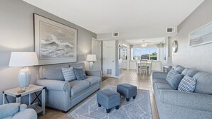Un salon moderne avec des canapés gris, une table basse et un tapis. On aperçoit à l’arrière-plan un coin repas avec une table et des chaises.