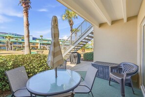 An outdoor patio with a round glass table and two chairs, a covered umbrella, and a staircase leading to a building with a green roof. - Seaspray Condos - 112A (Fort Walton Beach)