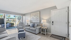 Un salon moderne avec un canapé gris, un petit pouf, un téléviseur et un balcon offrant une vue sur les immeubles.