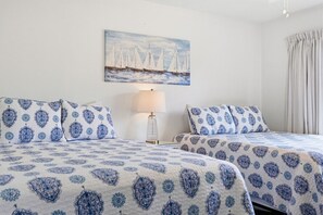 A hotel room with two beds, a nightstand, a lamp, and a painting of sailboats on the wall.