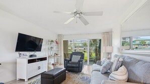Un salon équipé d’un téléviseur à écran plat, d’un ventilateur de plafond, d’un canapé, d’un fauteuil et d’une fenêtre donnant sur un immeuble.
