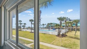 A view from a window overlooking a pool area with palm trees and a building in the background. - Seaspray Condos - 113A (Fort Walton Beach)