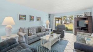 A living room with a sofa, armchairs, a coffee table, and a TV. There are framed pictures on the wall and a window with a view of a garden. - Seaspray Condos - 113A (Fort Walton Beach)