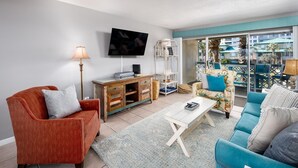 Un salon équipé d’un téléviseur, d’un canapé, d’un fauteuil, d’une table basse et offrant une vue sur une piscine et des palmiers à l’extérieur.