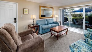 Un salon avec un fauteuil marron, un canapé bleu, une table basse en bois et un fauteuil à motifs. Un grand miroir est accroché au mur, et une porte est équipée d’un système de verrouillage à clavier.