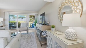 A modern living room with a sofa, a coffee table, a TV, and a decorative mirror. - Seaspray Condos - 115A (Fort Walton Beach)