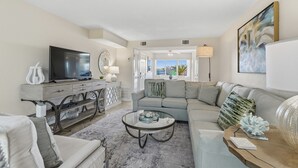 A modern living room with a grey sofa, a glass coffee table, and a TV on a wooden console. - Seaspray Condos - 115A (Fort Walton Beach)