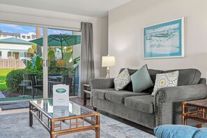 Une chambre d’hôtel avec un canapé gris, une table basse en verre et une vue sur un jardin et un immeuble, visible à travers une porte vitrée.