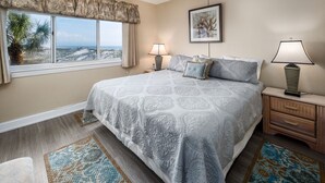 A bedroom with a large bed, two bedside lamps, a nightstand, a window with curtains, and a framed picture on the wall. - Seaspray Condos - 114D (Fort Walton Beach)