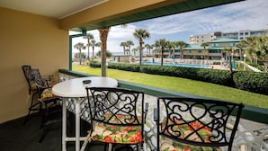A balcony with a table and chairs, overlooking a pool and palm trees. - Seaspray Condos - 114D (Fort Walton Beach)