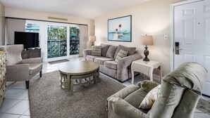 A living room with a sofa, armchair, coffee table, and a TV. There is a door with a sign above it, and a painting on the wall. - Seaspray Condos - 114D (Fort Walton Beach)