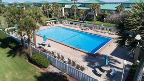 A swimming pool with lounge chairs and umbrellas, surrounded by palm trees and a building with a green roof.
