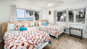 Une chambre avec deux lits, de grandes fenêtres donnant sur l’océan, un ventilateur de plafond et un plancher en bois.
