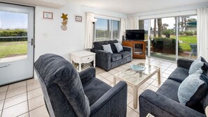 Un salon comprenant un canapé, des fauteuils, une table basse et un téléviseur. Une porte-fenêtre coulissante donne sur un balcon d’où l’on peut admirer un paysage verdoyant et un immeuble.