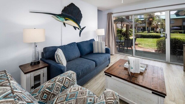 A modern hotel room with a blue sofa, patterned cushions, a wooden side table, and a large window offering a view of the outdoor area.