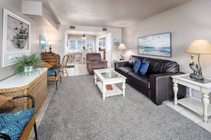 A living room with a sofa, coffee table, and chairs, a dining area with a table and chairs, and a kitchen in the background.