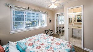 A bedroom with a bed, a window with blinds, a ceiling fan, and a bathroom with a mirror and sink. - Seaspray Condos - 204C (Fort Walton Beach)