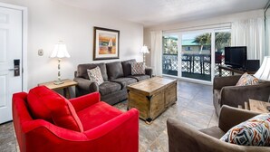 A living room with a red armchair, a sofa, a coffee table, and a TV. There is a painting on the wall and a balcony with a view. - Seaspray Condos - 204C (Fort Walton Beach)