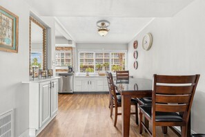 Una cocina con armarios blancos, una mesa de comedor de madera y sillas. Hay ventanas con persianas, una lámpara en el techo y relojes de pared.