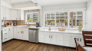 A kitchen with white cabinets, a dishwasher, a coffee maker, and a window with blinds overlooking a building. - Seaspray Condos - 204D (Fort Walton Beach)