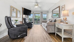 A spacious room with a ceiling fan, a flat-screen TV, a sofa, a desk, and a view through a glass door to an outdoor area. - Seaspray Condos - 204D (Fort Walton Beach)