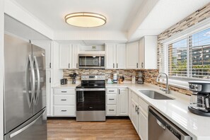 Una cocina moderna con armarios blancos, electrodomésticos de acero inoxidable, microondas integrado y un ventanal con persianas.