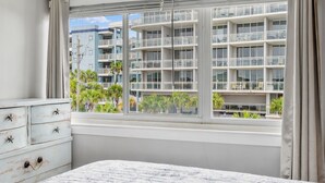 A bedroom with a large window, a bed, a dresser with star-shaped handles, and a view of a multi-story building with balconies. - Seaspray Condos - 204D (Fort Walton Beach)