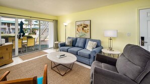 A living room with a blue sofa, a grey armchair, a round coffee table, and a small side table with a lamp. There is a large window with a view of a building outside. - Seaspray Condos - 206A (Fort Walton Beach)