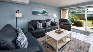 A living room with a black leather sofa, a white coffee table, and a blue wall with a wave painting. - Seaspray Condos - 207B (Fort Walton Beach)