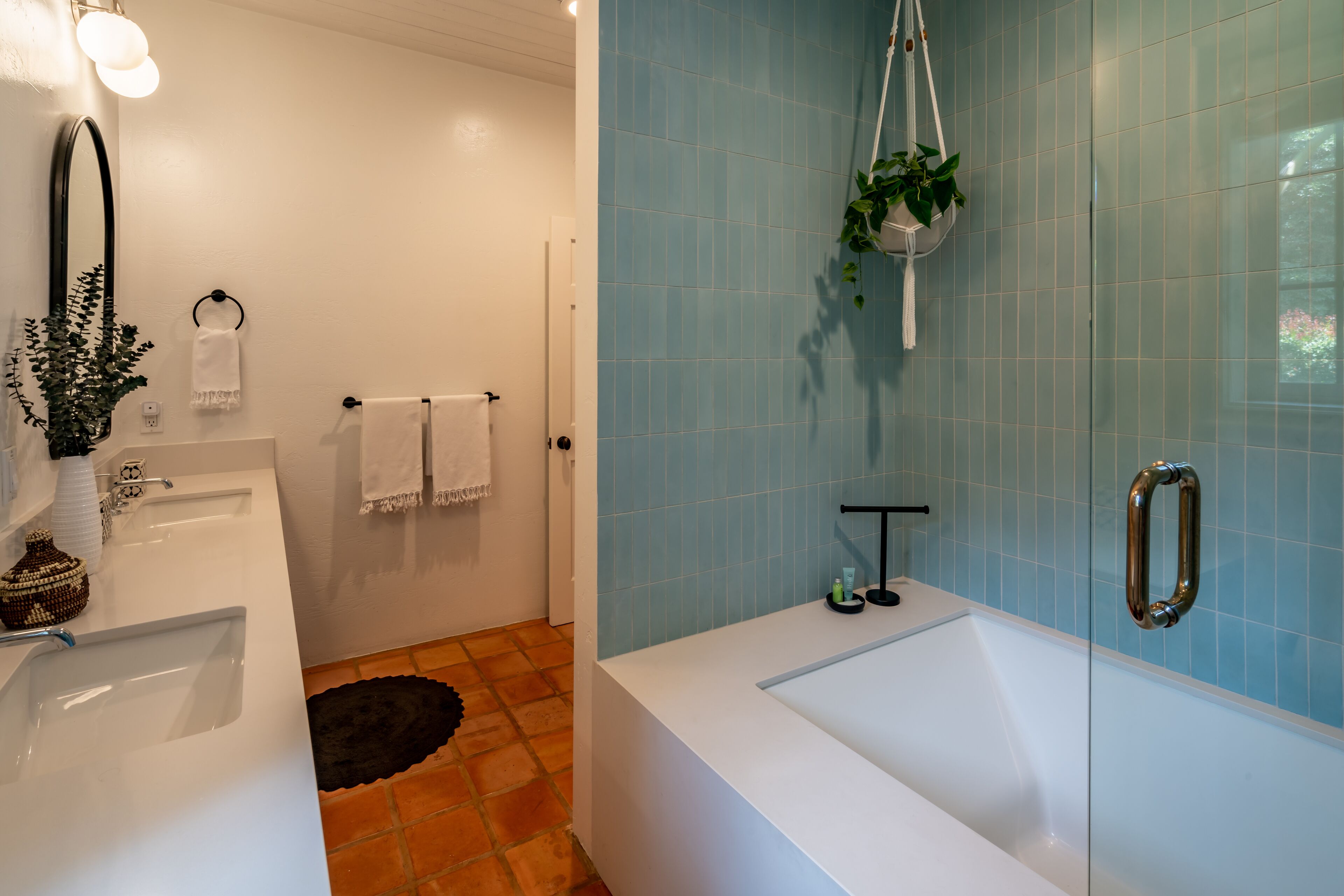 Une salle de bain avec un lavabo blanc, une baignoire et un espace douche. On y trouve un porte-serviettes avec des serviettes et une plante suspendue.