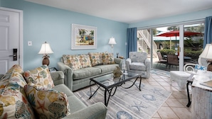 A living room with a sofa set, a coffee table, and a chair. There is a painting on the wall and a view of the outside through a glass door. - Seaspray Condos - 208B (Fort Walton Beach)