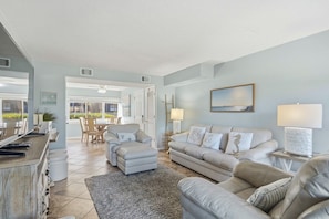 A spacious living room with a sofa set, a coffee table, and a TV. There is a dining area visible in the background.