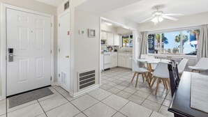 Une cuisine moderne avec des meubles blancs, un coin repas doté d’une table en verre et une vue sur les palmiers par la fenêtre.